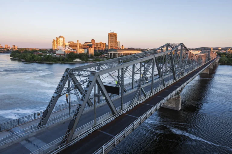 Een grote metalen tralie brug over het water met gebouwen van de skyline op de achtergrond bij schemering