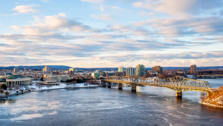 Stadsgezicht van Gatineau met gebouwen langs een deels bevroren rivier en een lange metalen spoorbrug bij zonsondergang