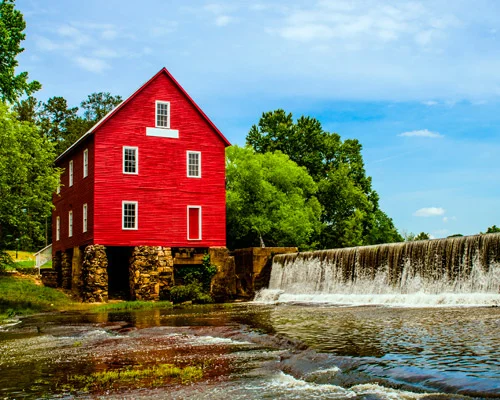 Historische rode houten watermolen op een stenen fundering naast een brede waterval, omringd door groene bomen in Georgia