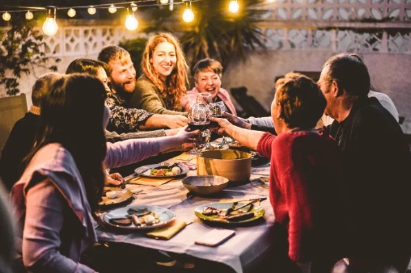 Groep volwassenen en kinderen klinkt met glazen aan een lange tafel buiten, verlicht door lichtsnoeren