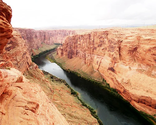 Smalle rivier met groenblauw water stroomt tussen hoge rode rotswanden van een diepe canyon onder een bewolkte lucht