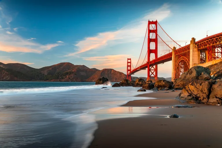 Golven spoelen over het zand terwijl de Golden Gate Bridge oplicht in het avondlicht op de achtergrond