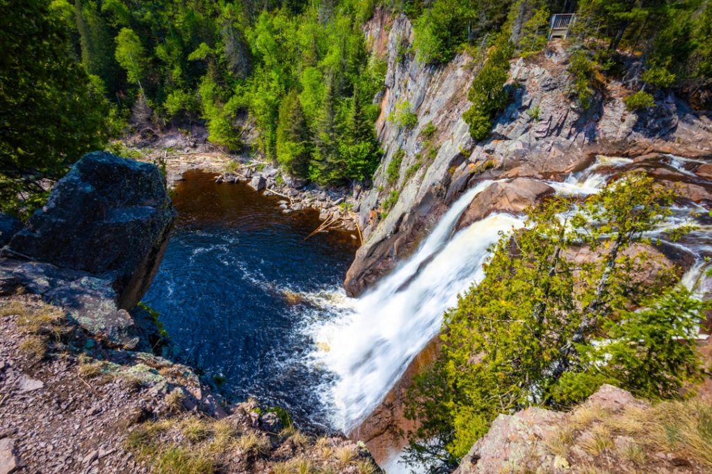 Luchtfoto van bovenaf op een waterval die in een donkere poel stort, omringd door rotsen en bos