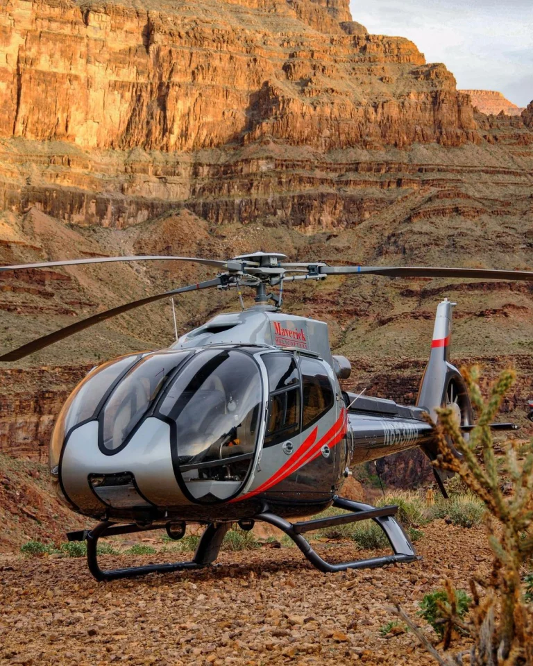 Een zilverkleurige helikopter van Maverick staat geland op een rotsachtige bodem tussen de steile, gelaagde rotswanden van de Grand Canyon