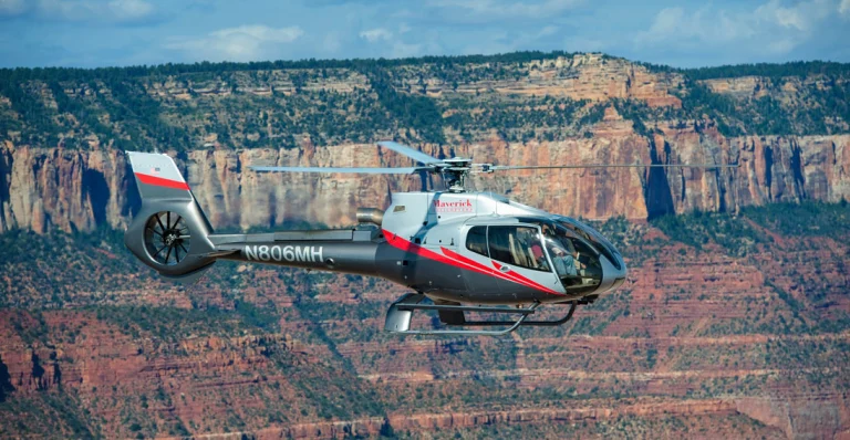Een zilverkleurige Maverick helikopter vliegt boven de gelaagde rotsformaties van de Grand Canyon