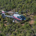Een witte en rode helikopter van Maverick vliegt boven een dicht bos met groene bomen en rode aarde bij de Grand Canyon