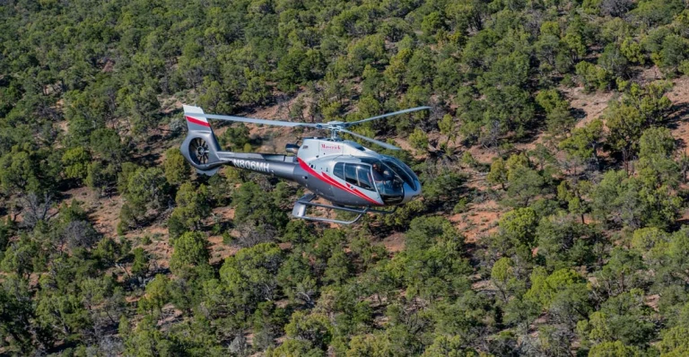 Een witte en rode helikopter van Maverick vliegt boven een dicht bos met groene bomen en rode aarde bij de Grand Canyon