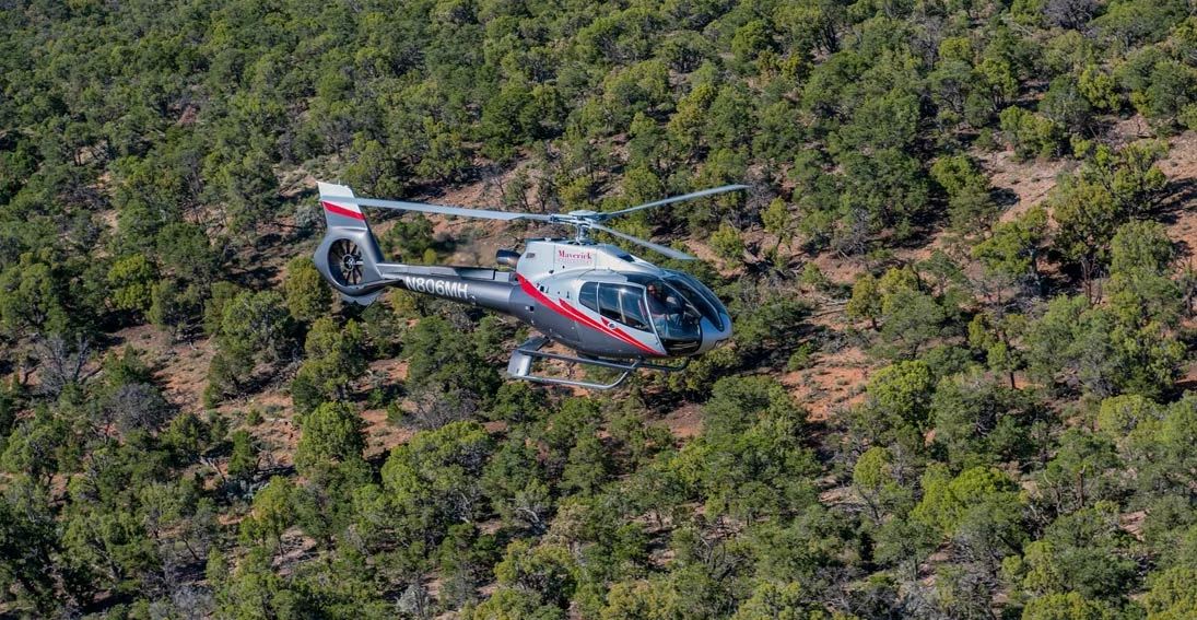 Een witte en rode helikopter van Maverick vliegt boven een dicht bos met groene bomen en rode aarde bij de Grand Canyon