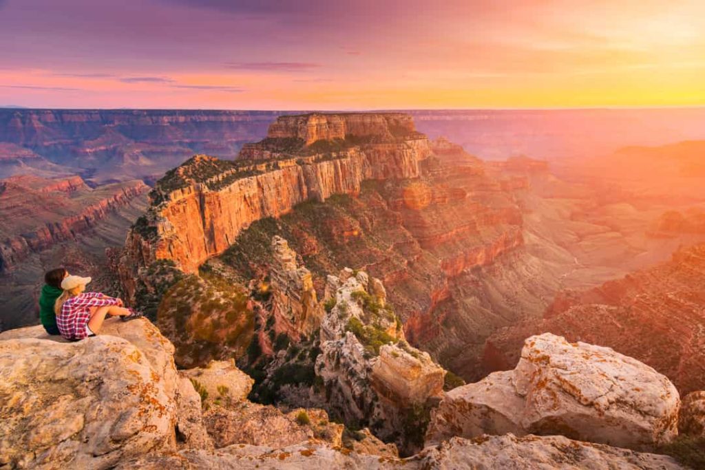 Twee wandelaars zitten op een rotspunt en kijken uit over de gelaagde kloofwanden van de Grand Canyon bij een gloeiende zonsondergang