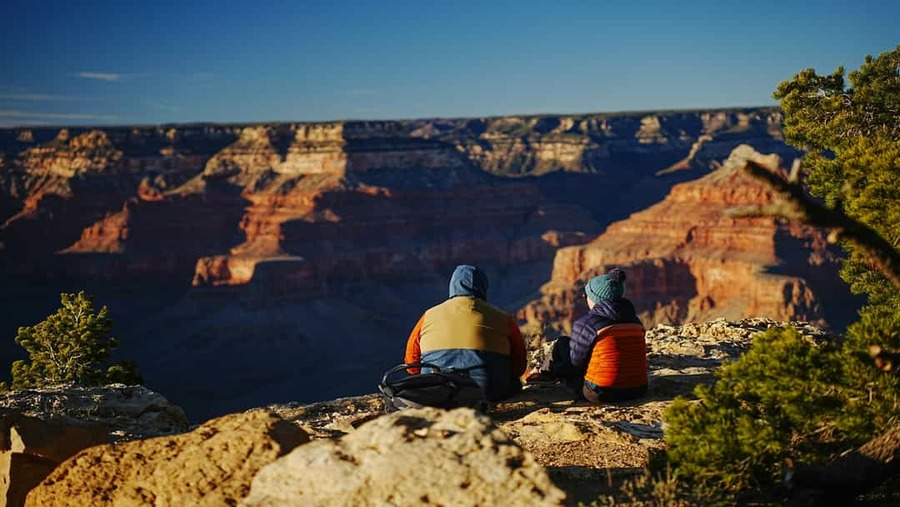 Twee mensen in warme kleding zitten op een rotsrand en kijken uit over de rood gekleurde rotsformaties van de Grand Canyon