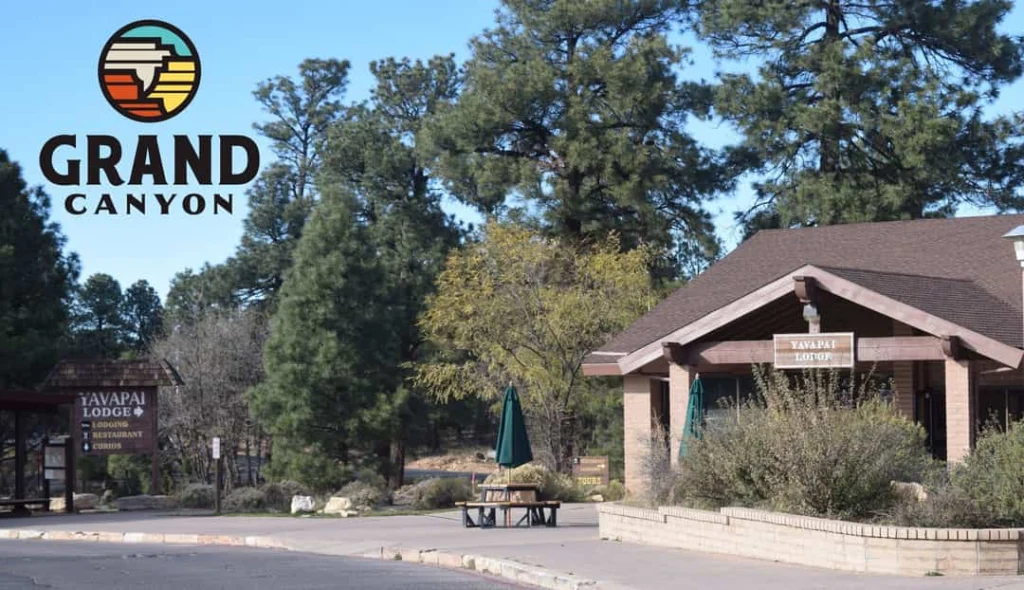 Gebouw van Yavapai Lodge met houten pergola en picknicktafel tussen dennenbomen met het Grand Canyon logo in beeld