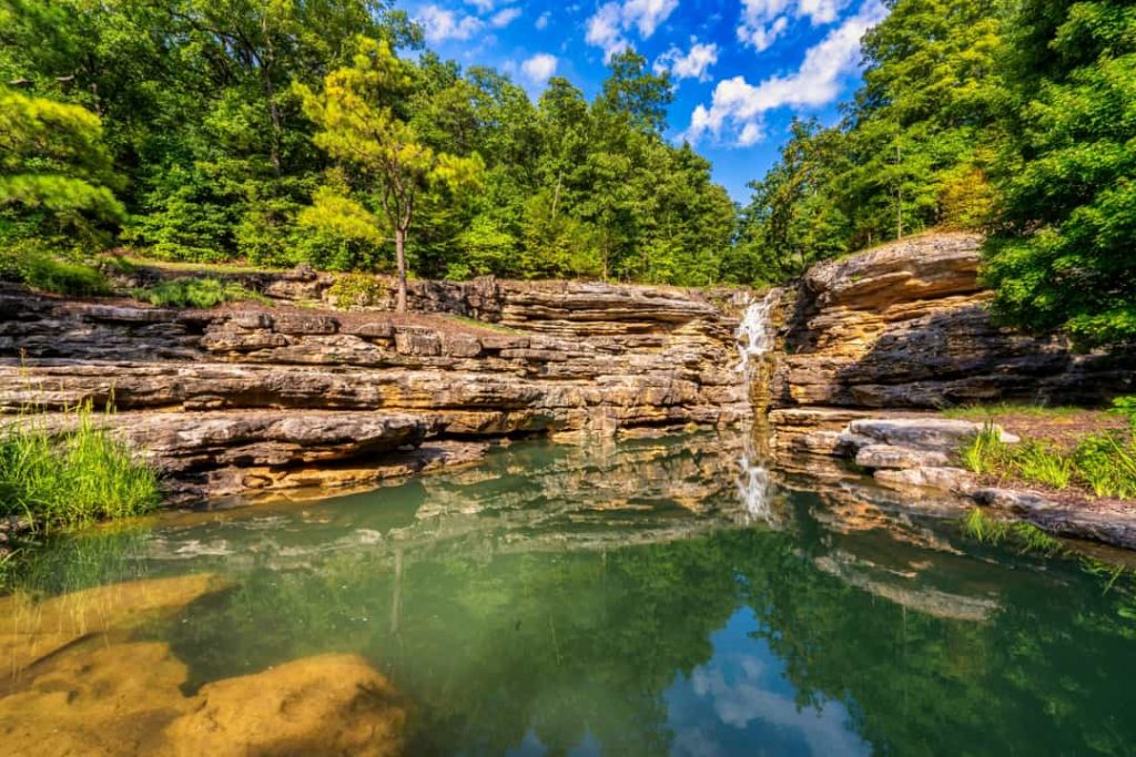 Brede waterval stroomt over een gelaagde rotsformatie in een groen bos, met een rustige poel water op de voorgrond in Missouri