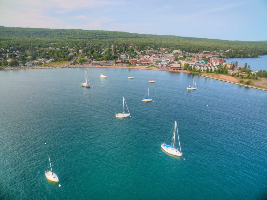 Luchtfoto van verspreide zeilboten op het blauwe water van Lake Superior, met het plaatsje Grand Marais op de achtergrond