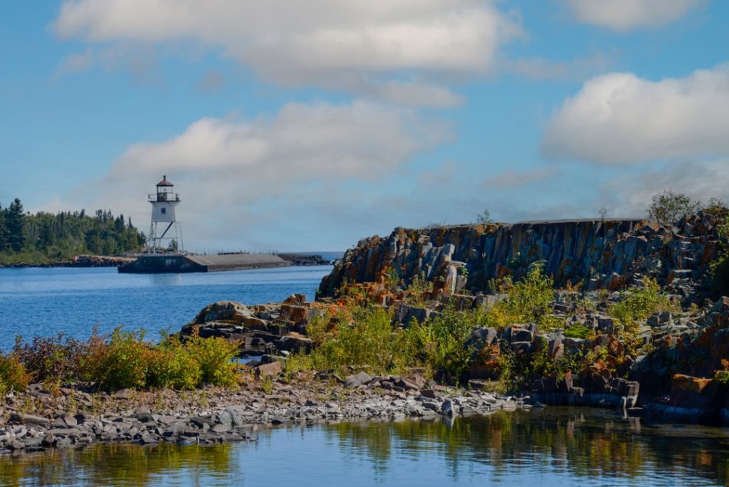 Witte vuurtoren op een verre pier gezien over rotsachtig water, met begroeide rotsen op de voorgrond in Grand Marais