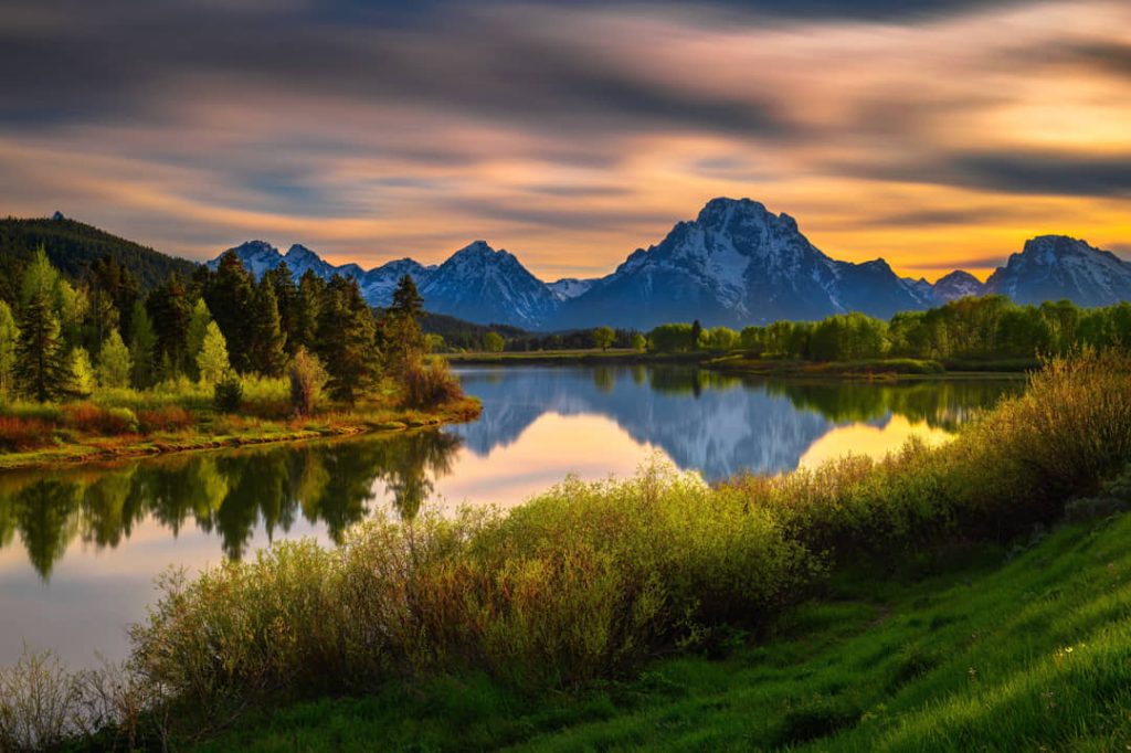 Kalm water van een rivierbocht dat de besneeuwde Teton bergketen en groene bomen weerspiegelt bij zonsondergang