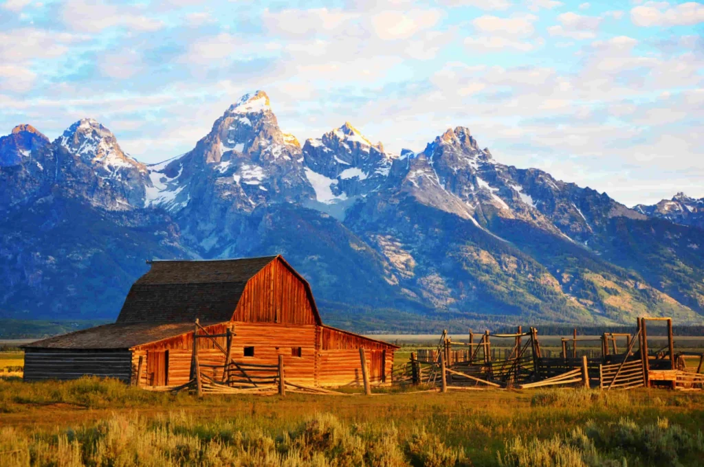 Houten schuur met veekraal in warm ochtendlicht, op de achtergrond de scherpe besneeuwde bergpieken van de Grand Teton tegen een blauwe lucht