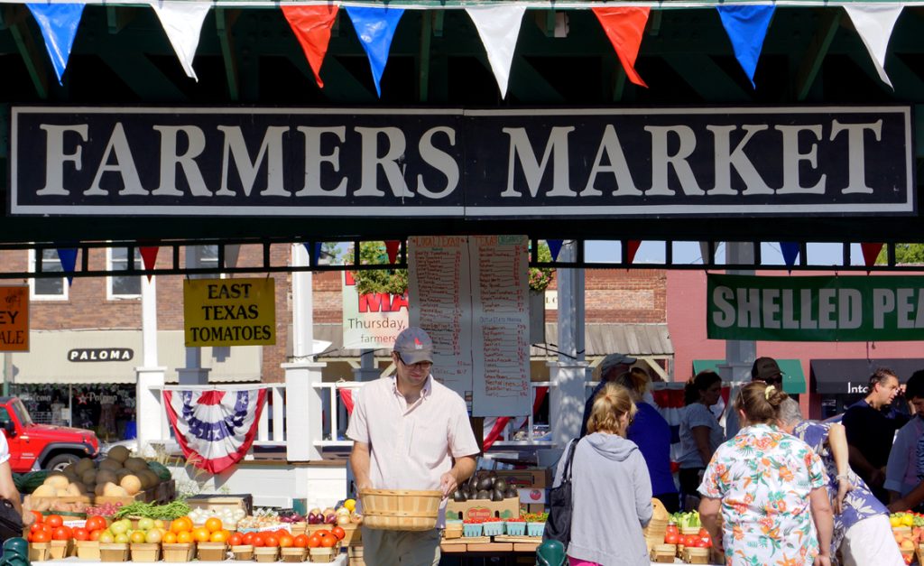Bezoekers bij een kraam met groente en fruit onder een bord met de tekst Farmers Market, versierd met vlaggetjes