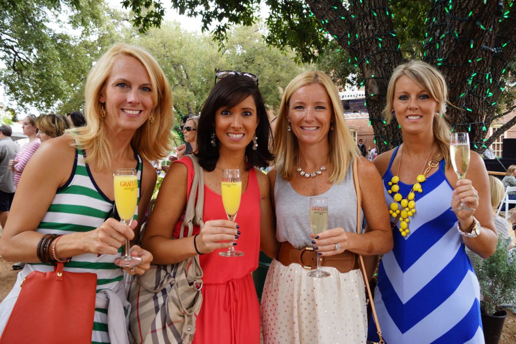 Vier lachende vrouwen heffen champagneglazen tijdens een buitenfeest onder bomen tijdens het wijnfestival GrapeFest in Grapevine