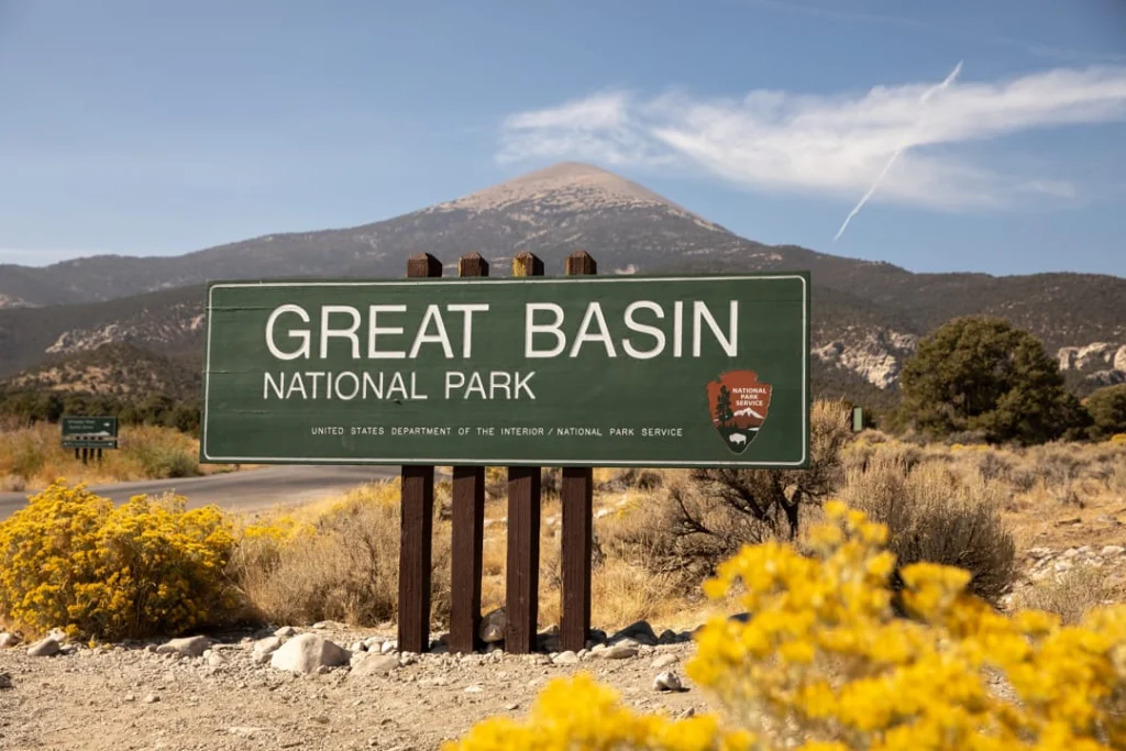 Groen bord met de tekst Great Basin National Park langs de weg, met bergen en gele struiken op de voorgrond