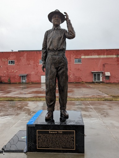 Bronzen standbeeld van een jonge man met hoed die groetend zijn hand opsteekt voor een rood bakstenen gebouw in Greenwood, Mississippi