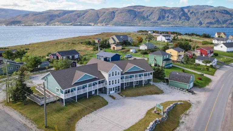 Groot huis met veranda's tussen kleinere woningen bij een baai, met bergen en een lange waterpartij op de achtergrond