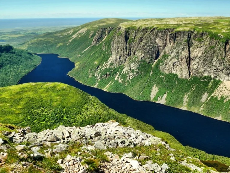 Blauw fjordmeer tussen groene beboste heuvels gezien vanaf een rotsachtig uitkijkpunt met stenen op de voorgrond in Gros Morne