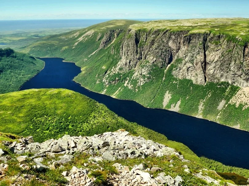 Blauw fjordmeer tussen groene beboste heuvels gezien vanaf een rotsachtig uitkijkpunt met stenen op de voorgrond in Gros Morne