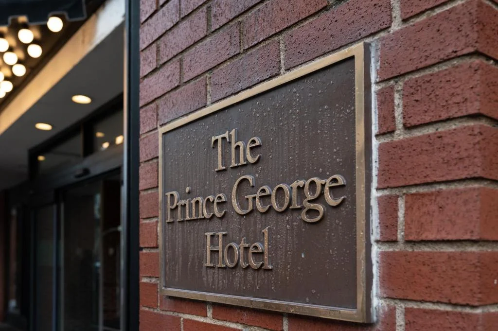 Bronzen plaquette met de tekst The Prince George Hotel bevestigd op een bakstenen gevel naast de ingang
