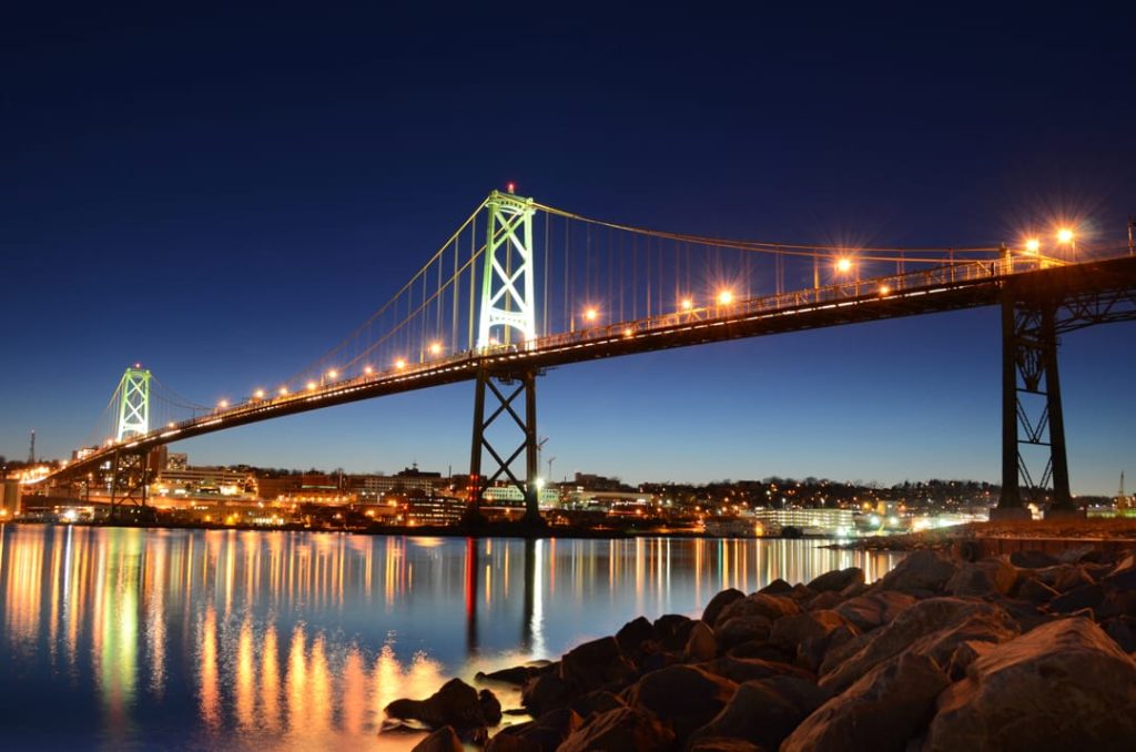 Grote verlichte hangbrug overspant het water met stenen op de voorgrond en de skyline van Halifax op de achtergrond