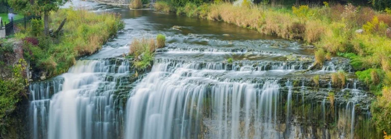 Brede waterval met meerdere stromen water die over een gelaagde rotswand vallen, omgeven door groen en gras bij Hamilton
