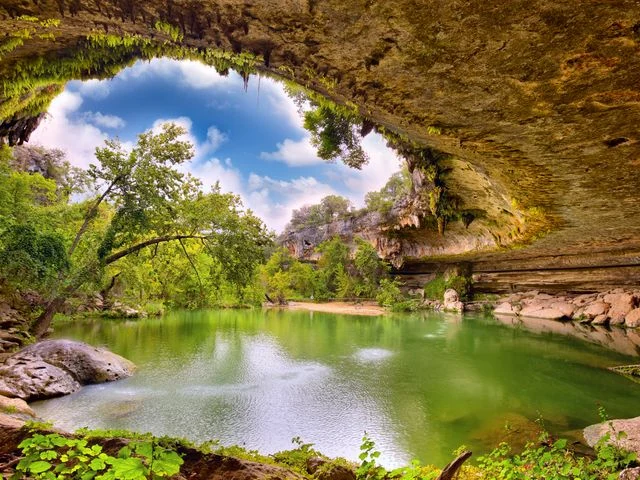 Groen water van een natuurlijk zwembad onder een overhangende rotsformatie begroeid met mos en bomen bij Hamilton Pool