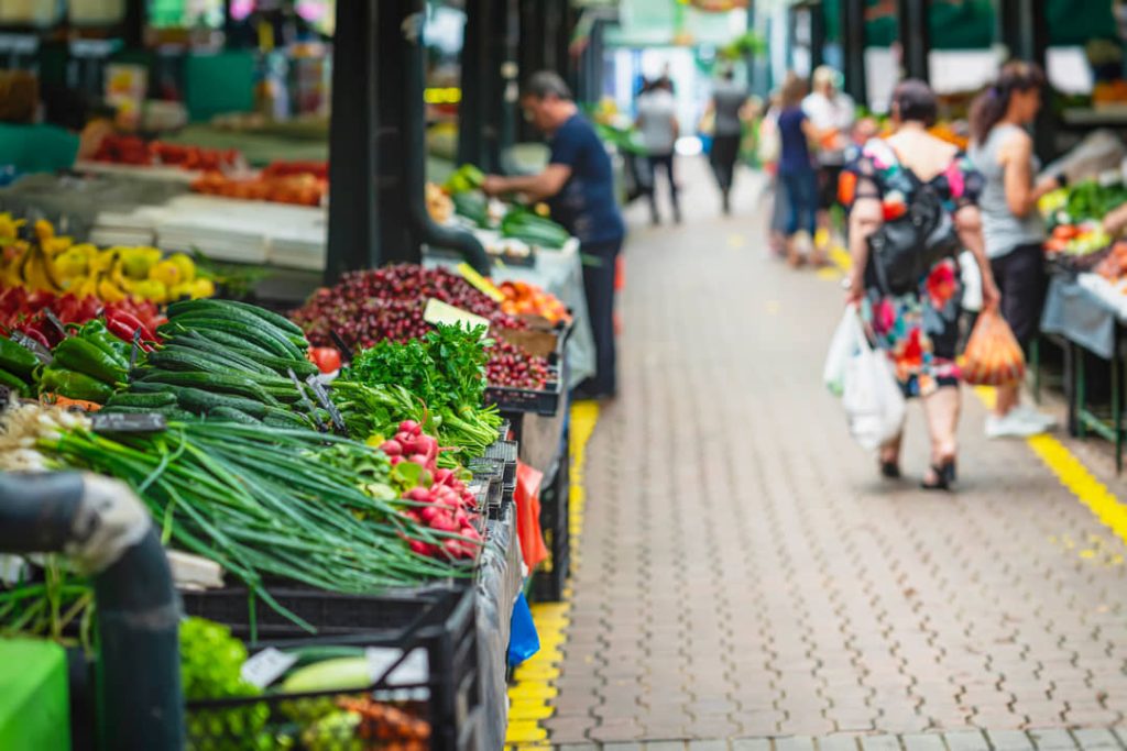 Kraam vol verse groenten zoals paprika's, komkommers en radijsjes op een drukke buurtmarkt met winkelende mensen