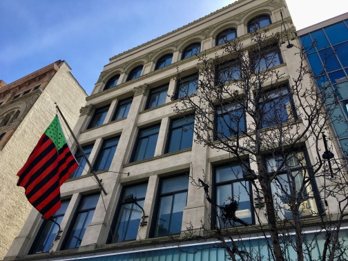 Hoge stenen gevel met ronde raamboogjes en een rood zwart groene vlag aan een vlaggenmast in Harlem