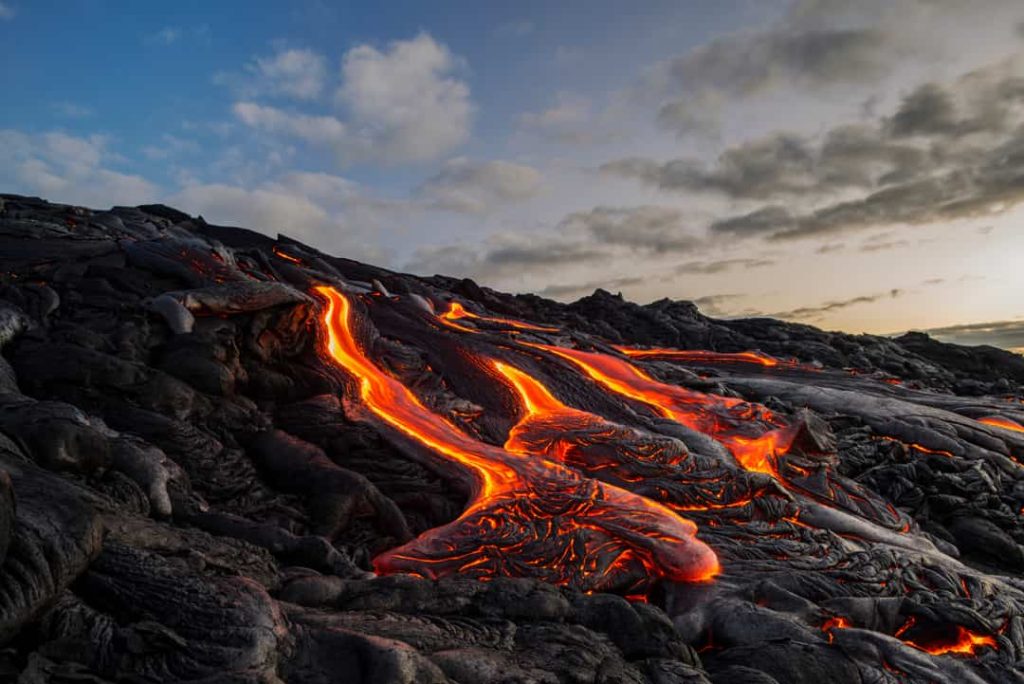 Oranje gloeiende lava die langzaam over afgekoeld zwart lavagesteente stroomt onder een schemerige avondlucht op Hawaii Groot Eiland