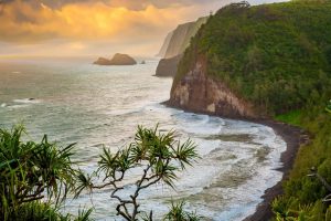Steile groene kliffen langs een zwart lavastrand met brekende golven onder een gouden avondlucht op Hawaii Groot Eiland