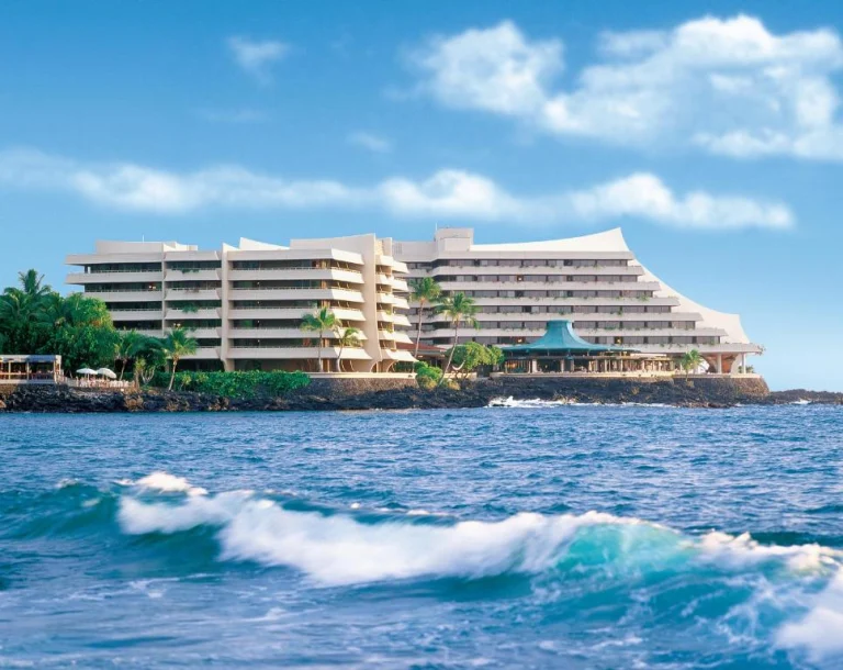 Een groot wit hotelcomplex met palmbomen staat op een rotsachtige kustlijn op Big Island, Hawaii, terwijl golven op de voorgrond breken