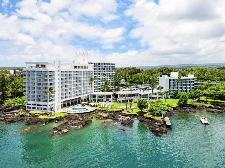 Luchtfoto van een wit hotelcomplex op Big Island, Hawaii, met zwembad en tuin naast een rotsachtige kustlijn en turquoise water