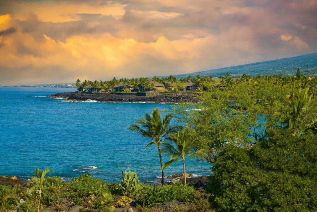 Resort met rieten daken langs de kust met palmbomen en een gouden wolkenlucht bij zonsondergang op Hawaii