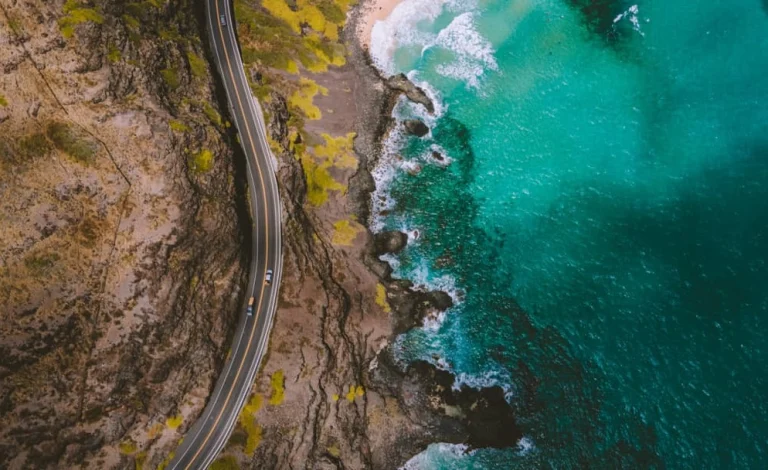 Luchtfoto van een kronkelende kustweg op Hawaii met rotsachtige kliffen en helderblauwe golven die breken op de kust