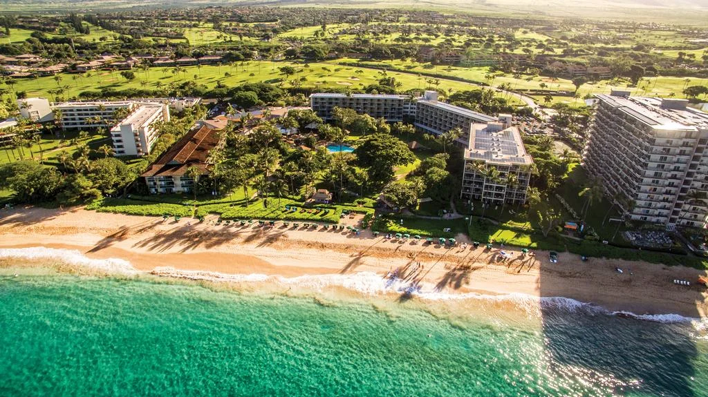 Luchtfoto van een kustlijn op Maui met meerdere hotels tussen groen gelegen aan een zandstrand met turquoise zee