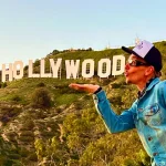 Een man in spijkerjasje en pet blaast een kus in de richting van het iconische Hollywood-bord op de heuvel