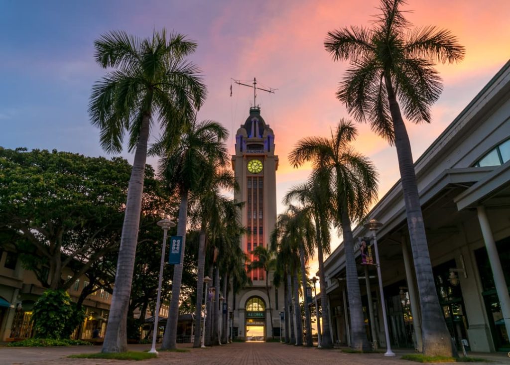 Verlichte klokkentoren van de Aloha Tower tussen hoge palmbomen tegen een roze en oranje avondlucht in Honolulu