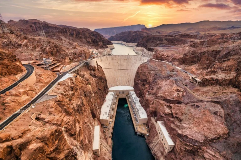 Luchtfoto van een kronkelende weg die afdaalt naar Hoover Dam, met een gouden zonsondergang boven de bergen