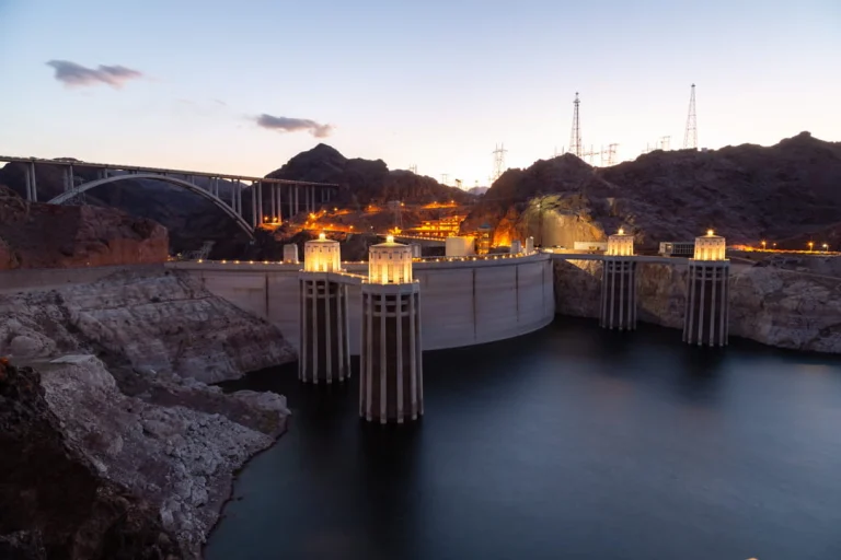 Hoover Dam met verlichte inlaattorens bij schemering, met de boogbrug en verlichte heuvels op de achtergrond