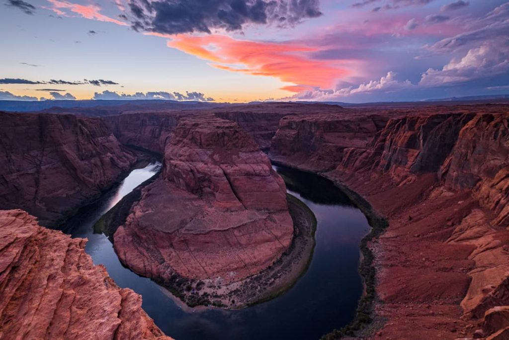 De hoefijzervormige bocht van de Colorado River tussen steile rode rotswanden, met een roze en paarse wolkenlucht erboven