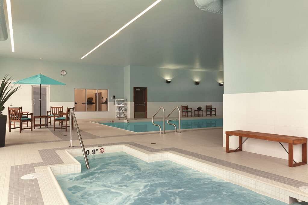 Overdekt zwembad met een bubbelende whirlpool op de voorgrond, houten loungestoelen, een blauwe parasol en een lang zwembad op de achtergrond