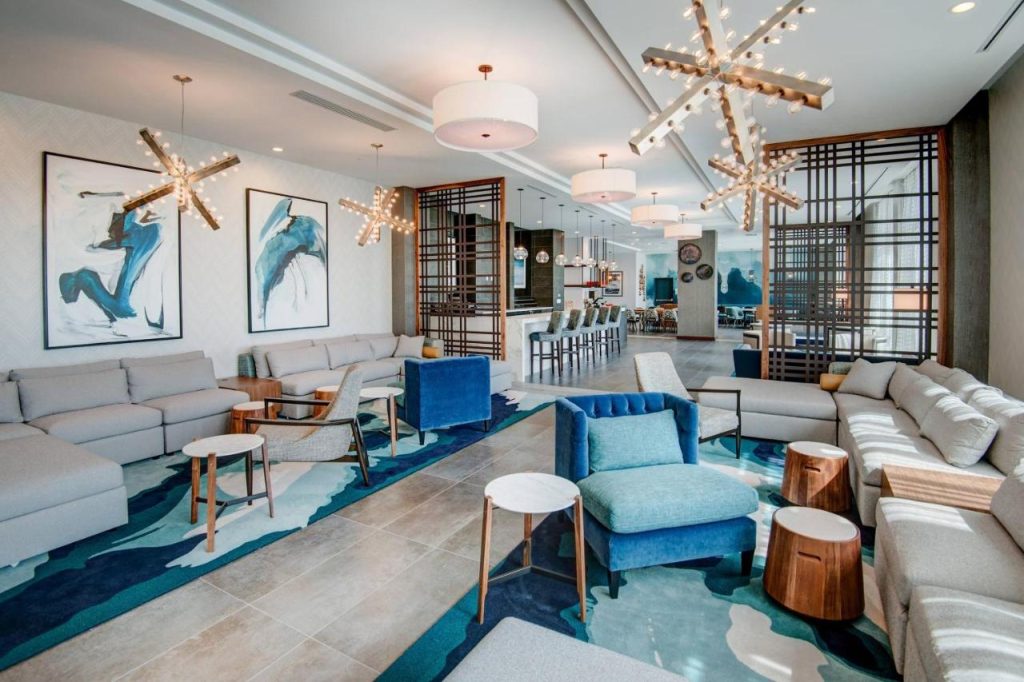 Hotellounge met grijze banken, een blauwe fauteuil en abstracte schilderijen aan de muur onder stervormige hanglampen