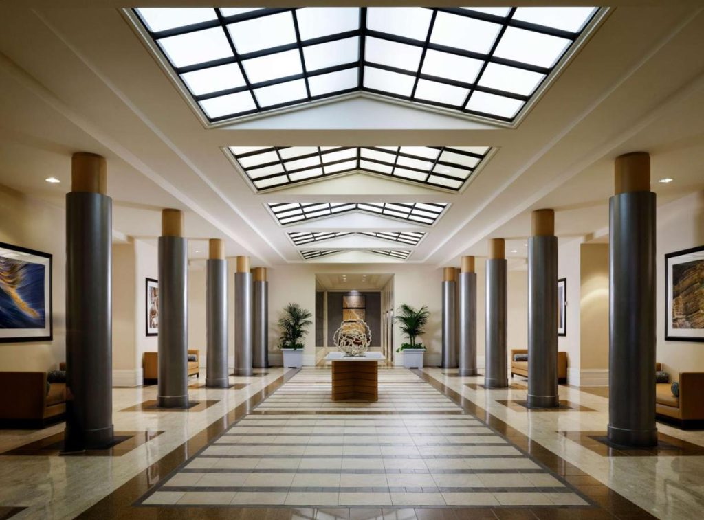 Lange hotellobby met zuilen, een marmeren vloer in schaakbordpatroon en driehoekige glazen lichtkoepels in het plafond