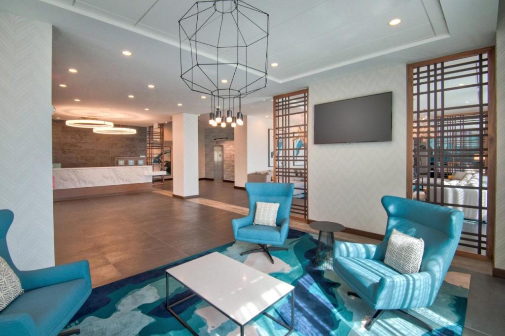 Hotellobby met turquoise fauteuils rond een salontafel, een televisie aan de muur en een geometrische hanglamp