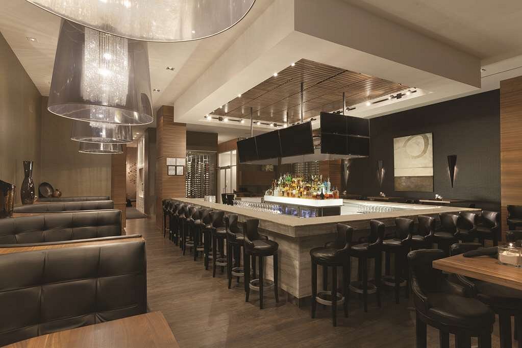 Lange bar met verlichte flessenrekken en leren barkrukken, met zwarte lederen booths en tafels ernaast in een modern verlicht hotelrestaurant